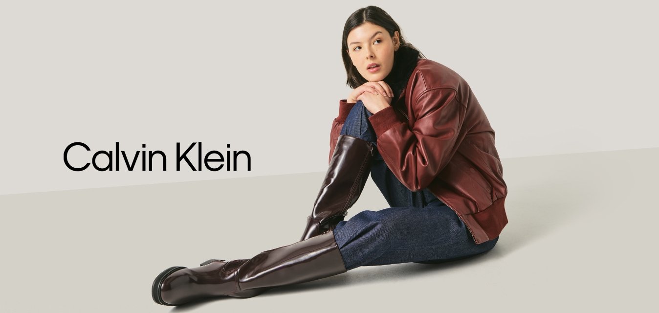 Mladá žena sedící na podlaze, oblečená v tmavě červené kožené bundě, tmavě modrých kalhotách a vysokých tmavě hnědých botách na jednoduchém světlém pozadí.