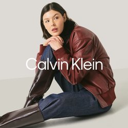 Mladá žena sedící na podlaze, oblečená v tmavě červené kožené bundě, tmavě modrých kalhotách a vysokých tmavě hnědých botách na jednoduchém světlém pozadí.