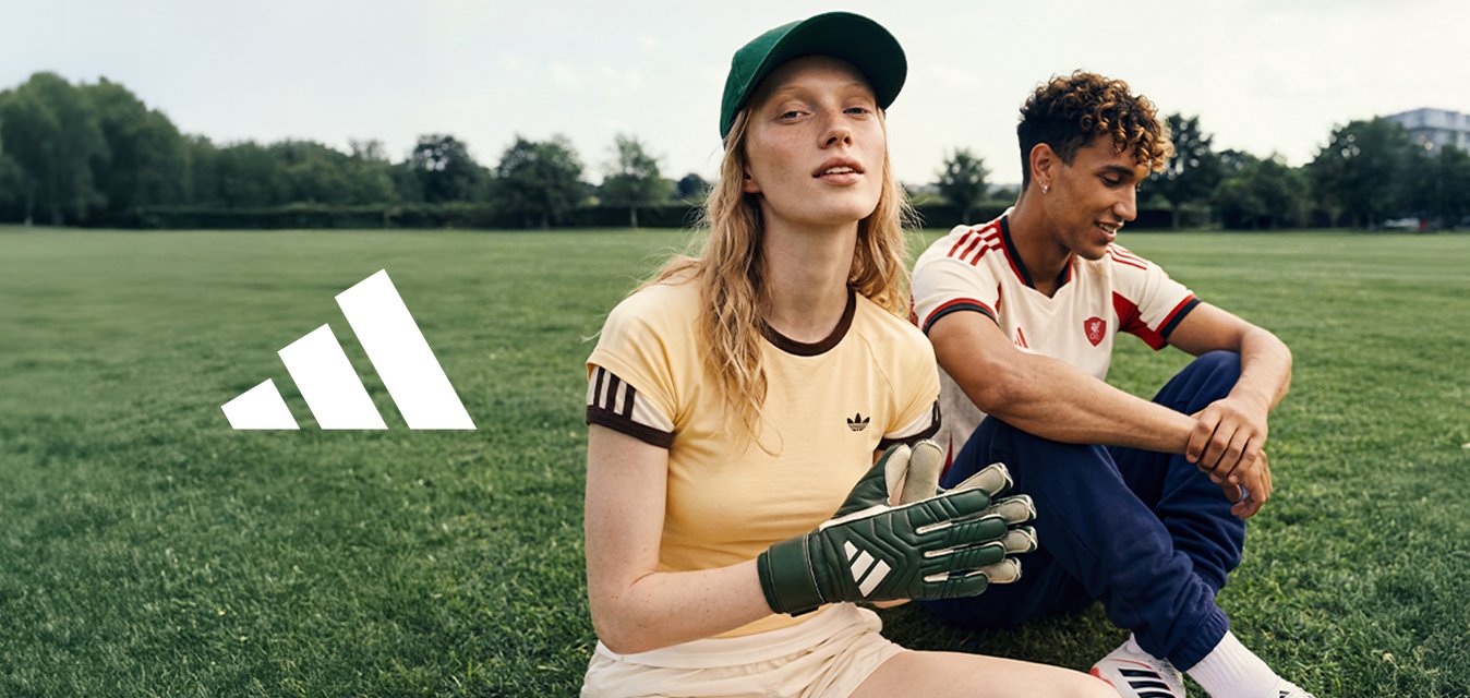 Dva mladí lidé v sportovním oblečení sedí na trávě, jeden má na sobě brankářské rukavice a čepici, druhý má na sobě fotbalový dres, venku.