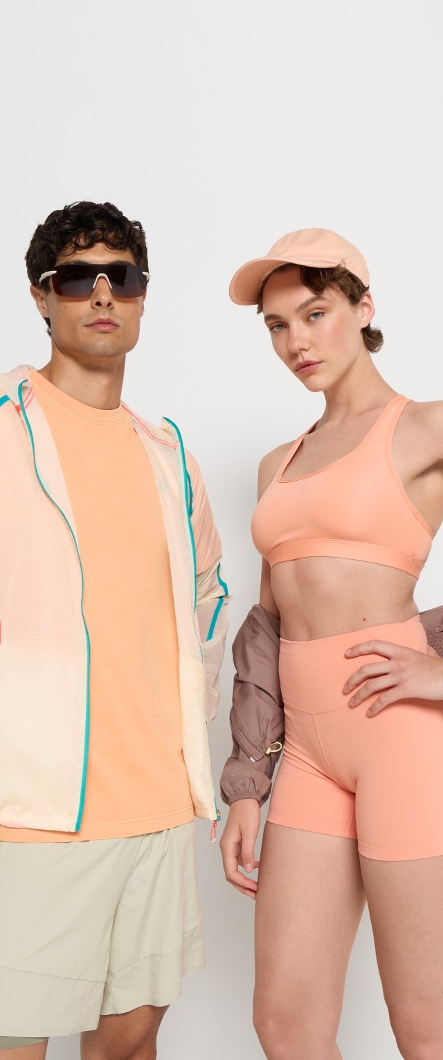 Due modelle indossano abbigliamento sportivo: una indossa un reggiseno sportivo arancione chiaro e pantaloni corti, mentre l'altra indossa una giacca leggera con accenti turchesi sopra una maglietta arancione.