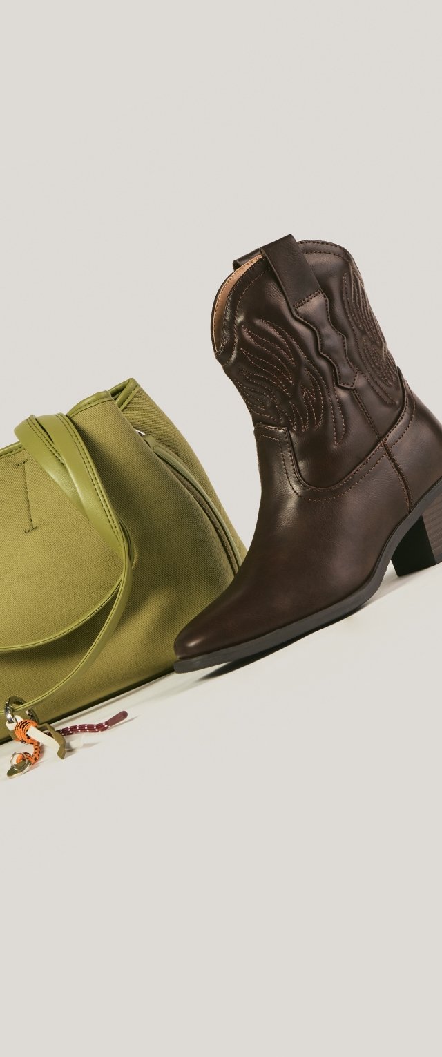 Bota de vaquero de cuero marrón oscuro con costuras decorativas junto a un bolso de tela verde oliva con correa para el hombro y llavero.