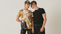 Zwei junge Erwachsene tragen Designerkleidung; einer in einem gemusterten gold-weißen Hemd mit Kappe und kleiner schwarzer Handtasche, der andere in einem schwarzen Polo mit gebrandetem Kragen.