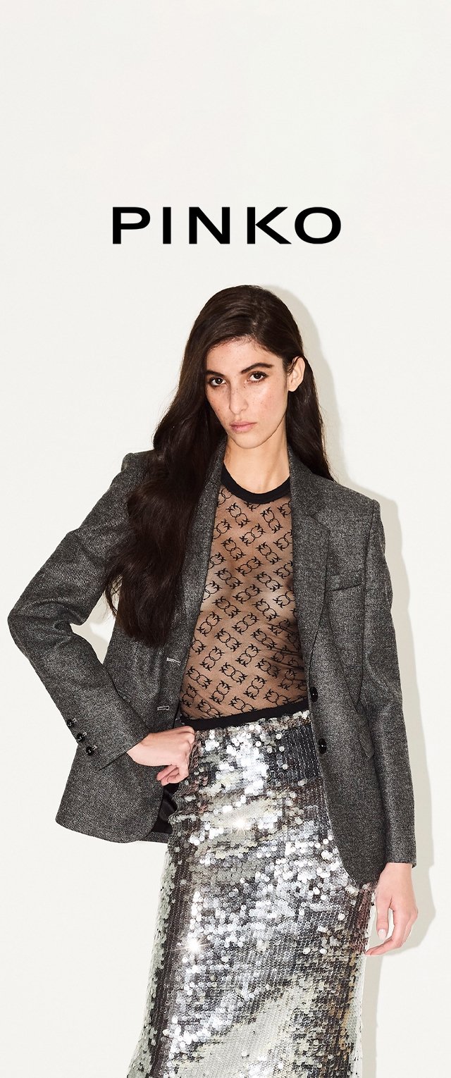 Blazer gris ajustado sobre una camiseta de malla negra con un patrón de logotipos repetido, combinado con una falda de lentejuelas plateadas brillantes.