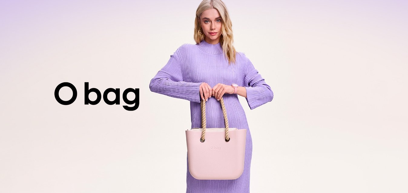 Žena v dlouhých levandulových šatech držící světle růžovou kabelku s lněnými uchy, stojící na jednoduchém světlém pozadí.