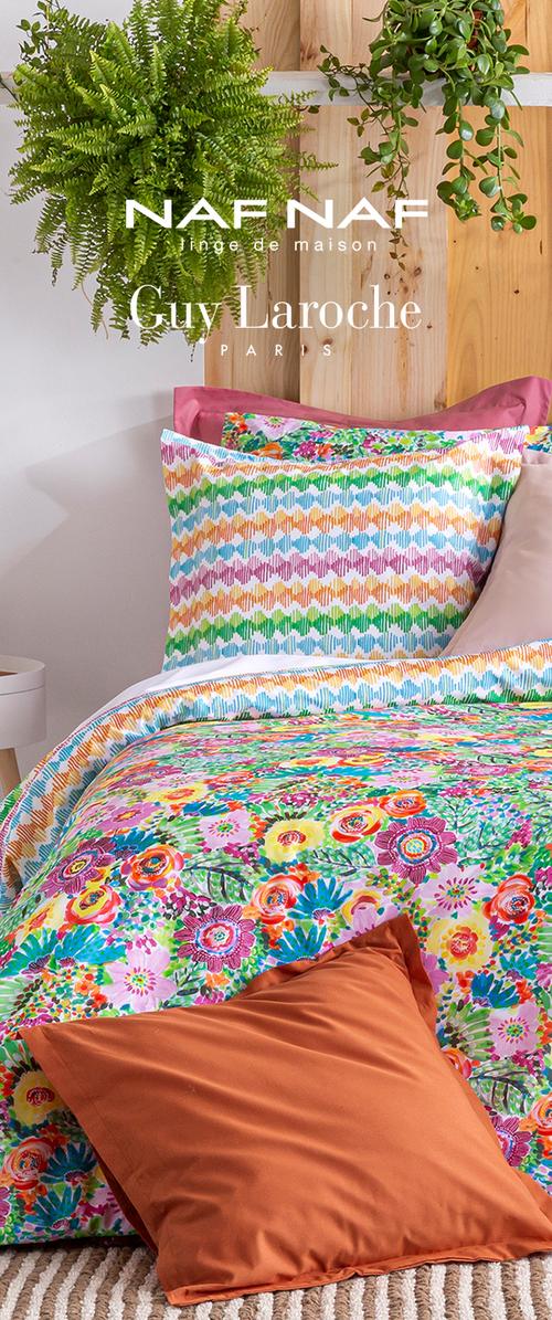 Colcha floral colorida con cojines estampados multicolor, cojín naranja sobre una alfombra texturizada, cabecero de madera y plantas verdes colgantes encima.