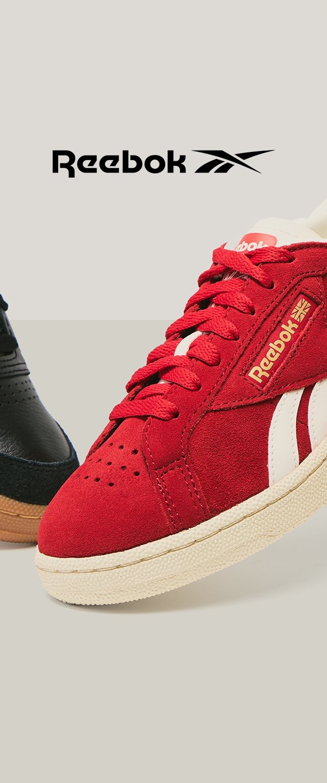 Czerwone zamszowe sneakersy z białymi akcentami, z okrągłymi sznurówkami, w stylu retro oraz teksturowaną podeszwą. Czarne sneakersy częściowo widoczne w tle.