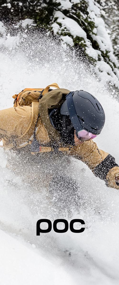 Escena de acción de esquí en nieve profunda; una persona lleva una chaqueta exterior marrón y un casco negro, con gafas rosas visibles y la nieve salpicando alrededor.