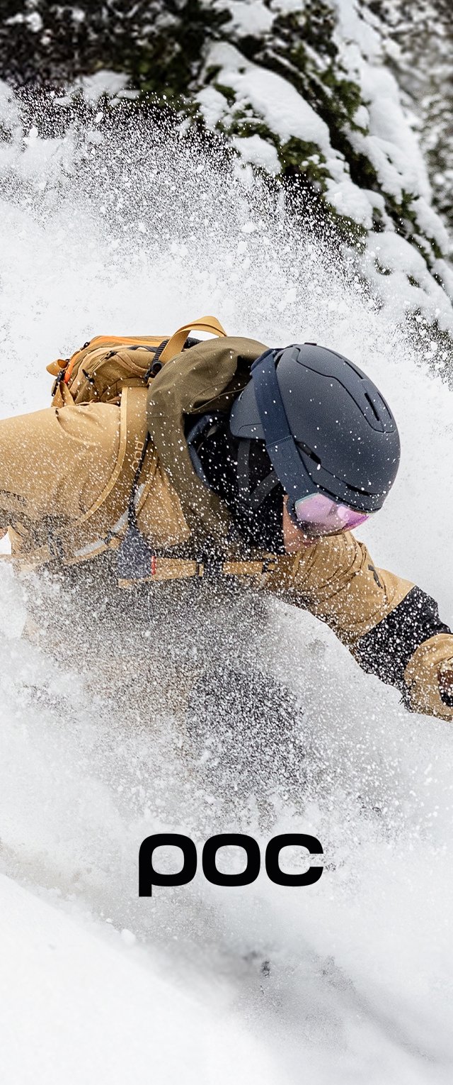 Escena de acción de esquí en nieve profunda; una persona lleva una chaqueta exterior marrón y un casco negro, con gafas rosas visibles y la nieve salpicando alrededor.