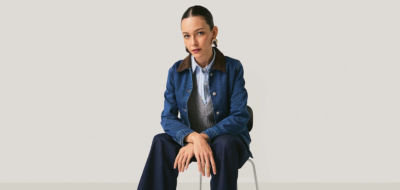 Džínová bunda s hnědým límcem, šedá vesta a světle modrá pruhovaná košile. Široké tmavé džíny a hnědé šněrovací boty. Sedí na židli.