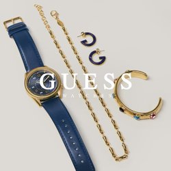 Hodinky s modrým koženým páskem, zlatý řetízkový náhrdelník, zlatý manžetový náramek s barevnými kameny a dva malé modro-zlaté kruhové náušnice na šedém pozadí.