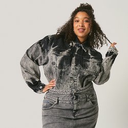 Žena s kudrnatými vlasy, oblečená v černošedé džínové bundě a sukni, stojící před jednoduchým světlým pozadím.