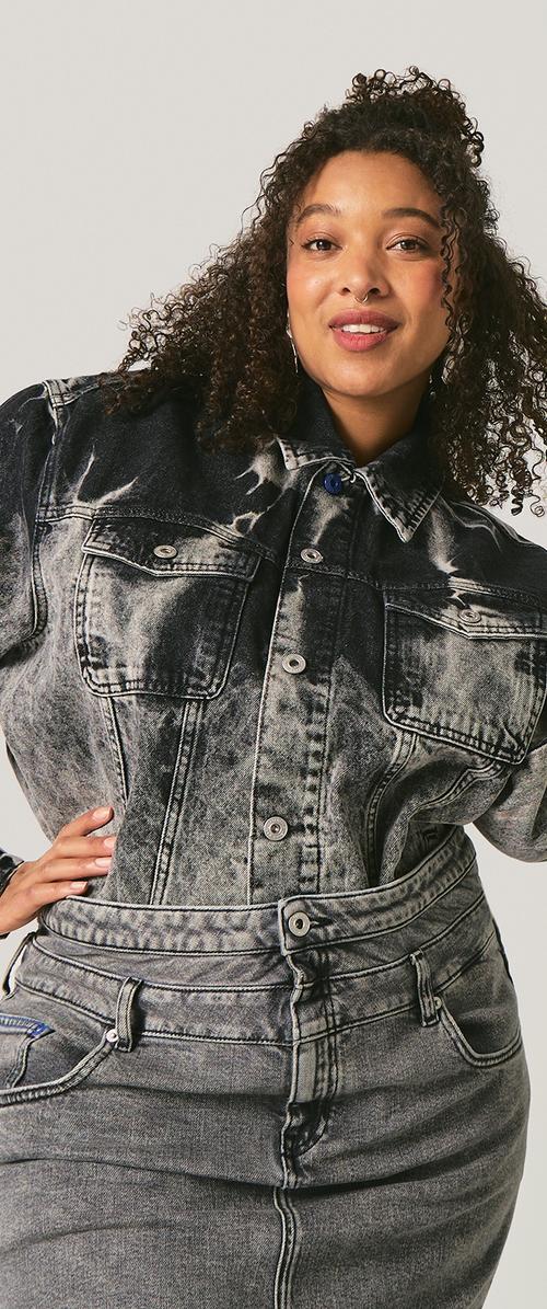 Mujer con cabello rizado sonriente que lleva una chaqueta vaquera negra blanqueada y jeans de mezclilla grises de talle alto, posando con una mano en la cadera.