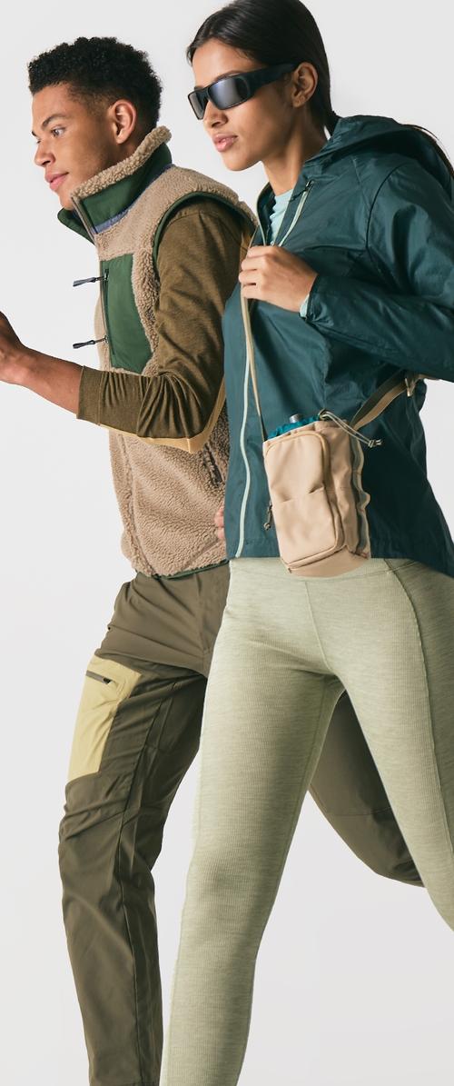 Deux individus marchent. L'un porte un gilet en polaire brun clair par-dessus des couches, tandis que l'autre est vêtu d'une veste à capuche verte et de leggings vert clair.