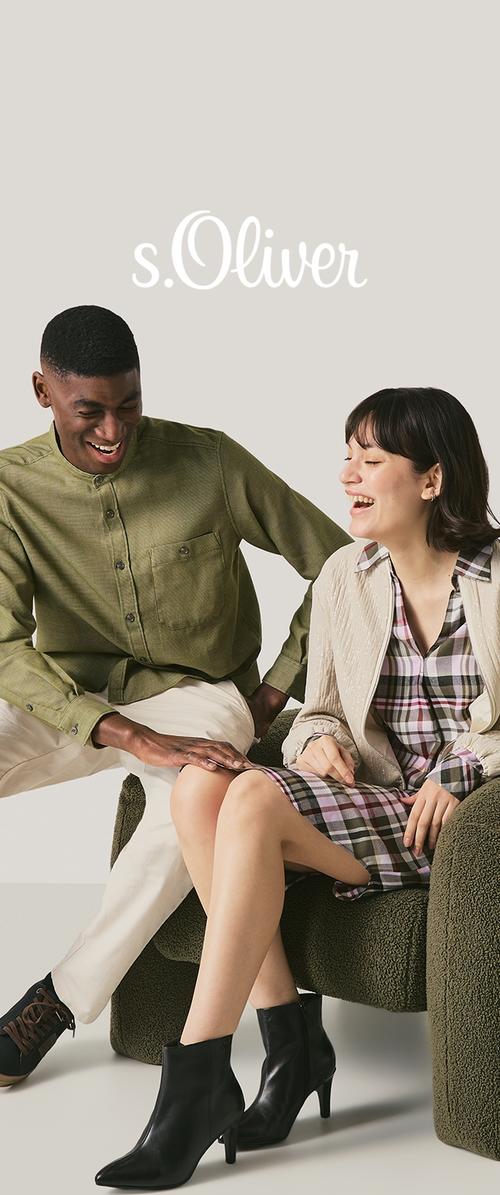 Un hombre con camisa verde y pantalones beige y una mujer con vestido de cuadros y chaqueta beige se sientan riendo en un sillón texturizado, con el logotipo de la marca arriba.