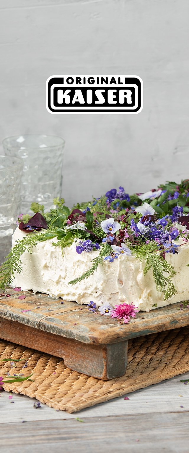 Gâteau crémeux et blanc garni de fleurs comestibles variées et de feuillage, présenté sur une planche en bois rustique, avec un tapis tissé en dessous.