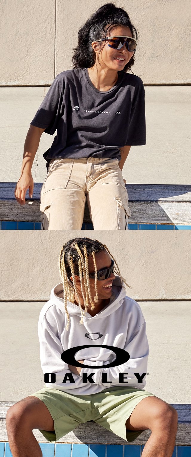 Dos personas sentadas en un borde. Una lleva una camiseta oscura y pantalones cortos beige; la segunda lleva una sudadera gris y pantalones cortos verdes, ambas con gafas de sol.