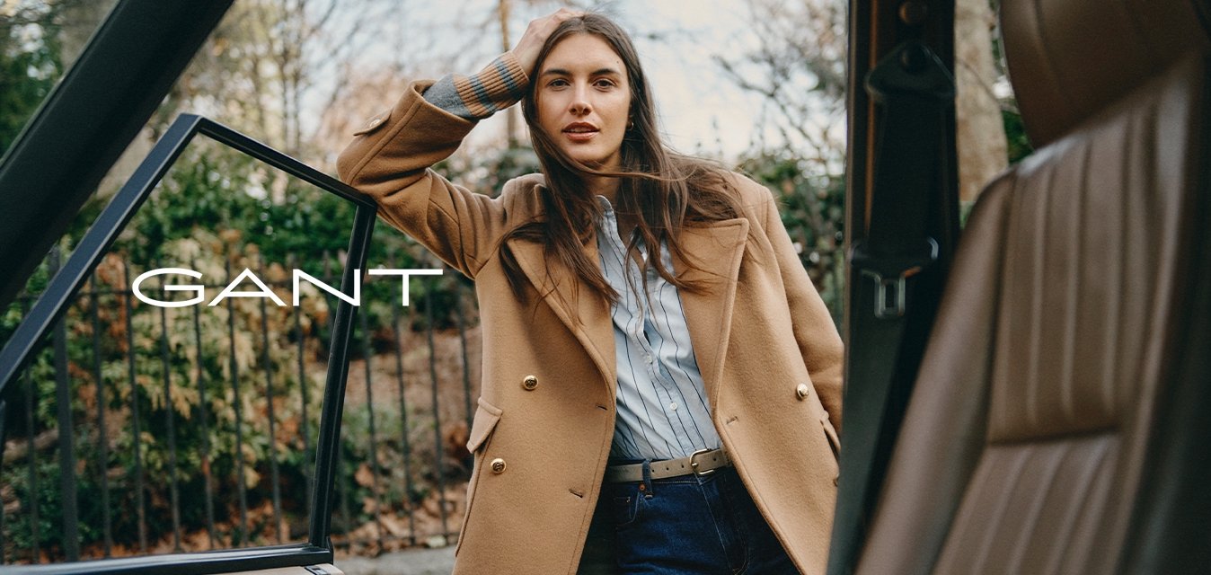 Hnědý vlněný kabát se zlatými knoflíky přes světle modrou pruhovanou košili, doplněné tmavými džínami. Stojí u otevřených dveří auta.