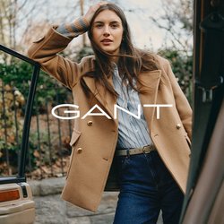 Hnědý vlněný kabát se zlatými knoflíky přes světle modrou pruhovanou košili, doplněné tmavými džínami. Stojí u otevřených dveří auta.