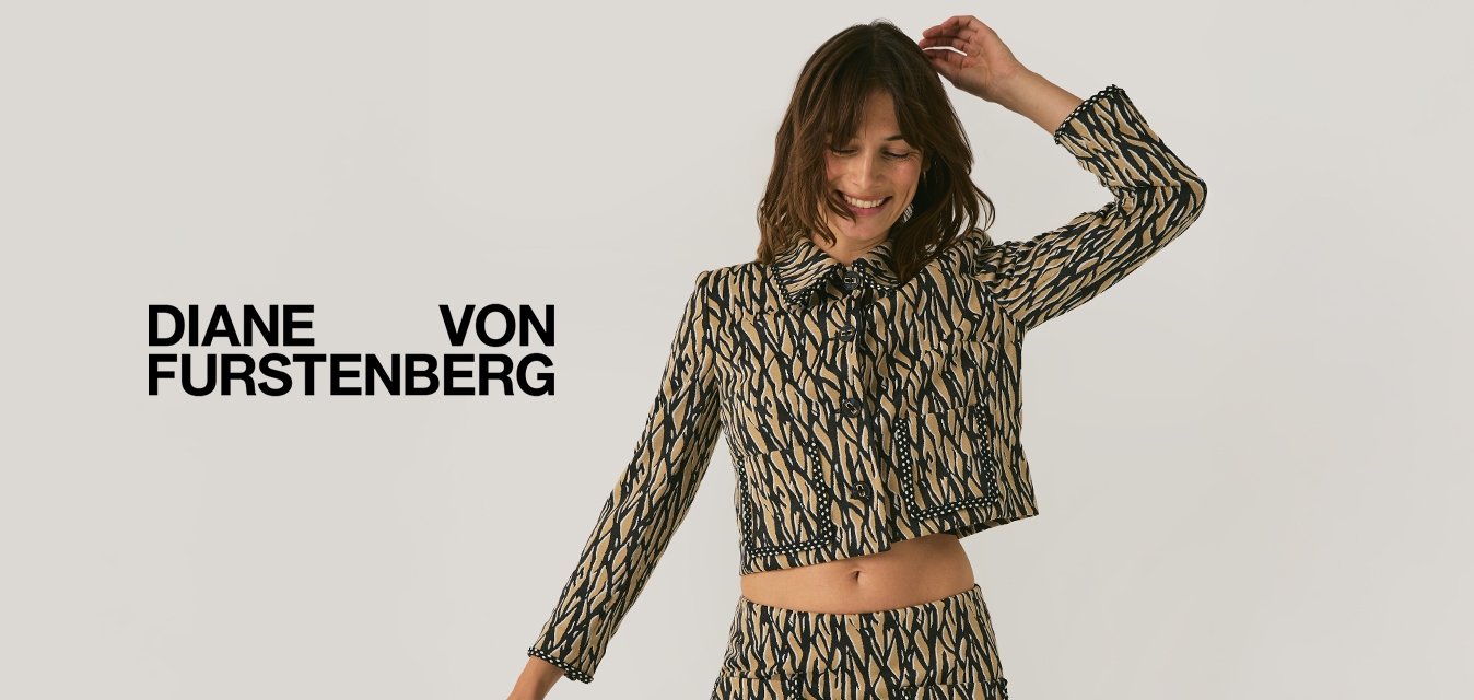 Žena v barevném krátkém saku a sukni s černými punčochami a botami pózuje s jednou zvednutou rukou a usmívá se.