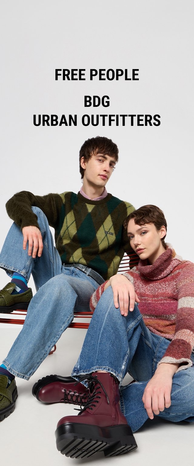 Two models sit against a red chair. One wears a green and black argyle sweater with blue jeans. The other sports a striped, red turtleneck and blue jeans. Both wear chunky boots.