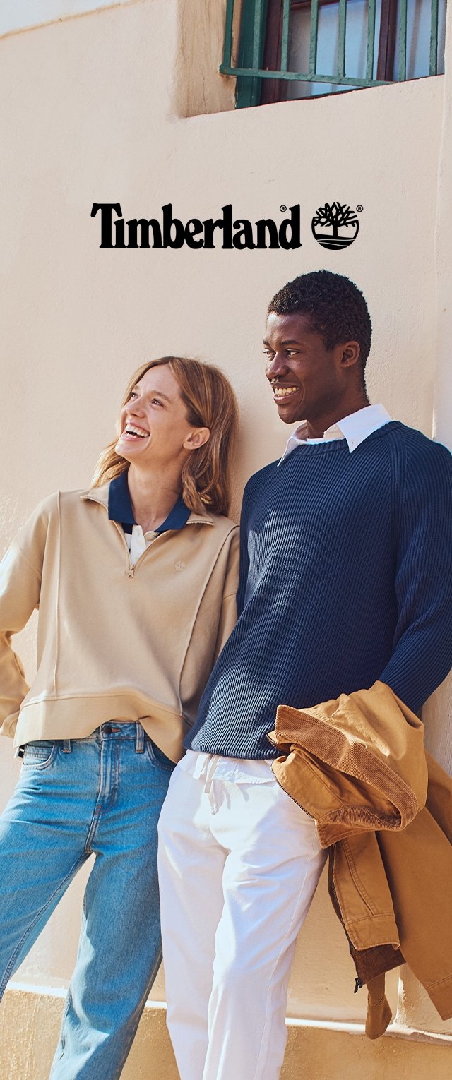 Due persone sono in piedi contro un muro chiaro. La donna indossa una maglia beige con zip sopra un colletto blu e dei jeans blu. L'uomo indossa un maglione blu navy a coste sopra una camicia bianca e pantaloni chiari. Tengono in mano una giacca marrone.
