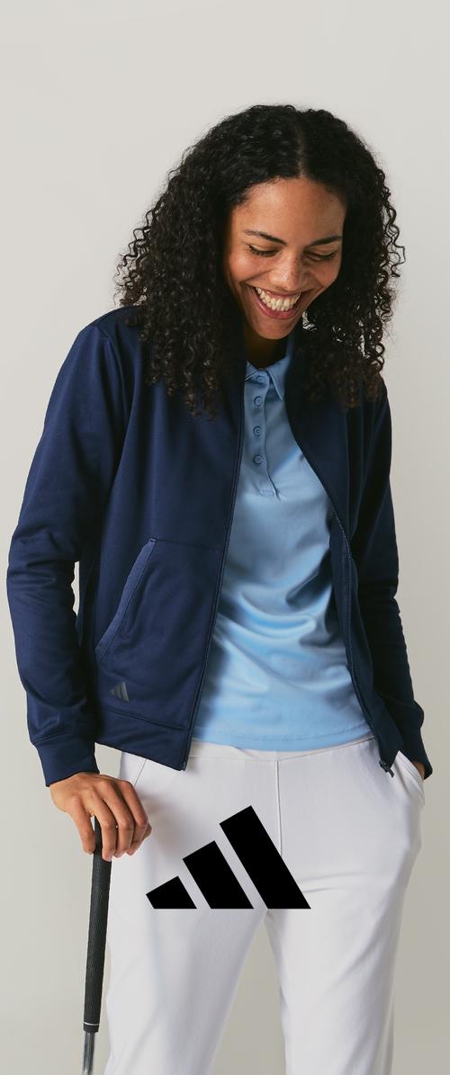 Veste zippée marine portée sur un polo bleu clair, associée à un pantalon blanc. Un club de golf est tenu dans une main ; logo Adidas visible sur le pantalon.