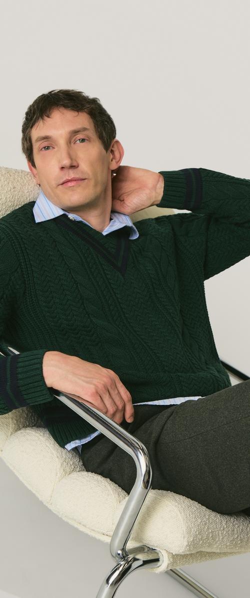 Maglione verde lavorato a maglia con scollo a V, indossato sopra una camicia celeste con colletto. Seduto su una sedia bianca con una texture, in una posa rilassata.