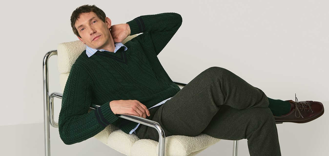 Muž se opírá v moderním bílém polstrovaném křesle s kovovým rámem, má na sobě zelený svetr, šedé kalhoty a hnědé boty na jednobarevném pozadí.