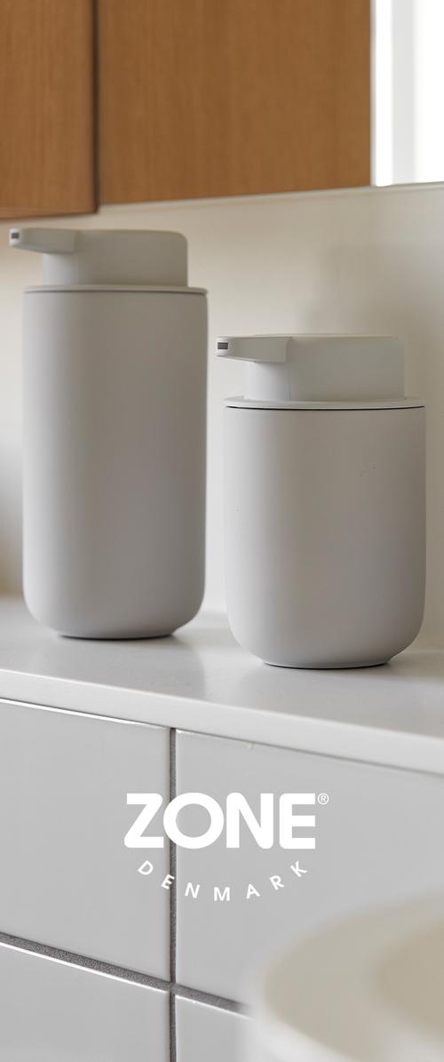 Dos dispensadores de cerámica blanca mate en dos tamaños con bombas en la parte superior. Formas lisas y cilíndricas con un diseño minimalista sobre una superficie de azulejos.