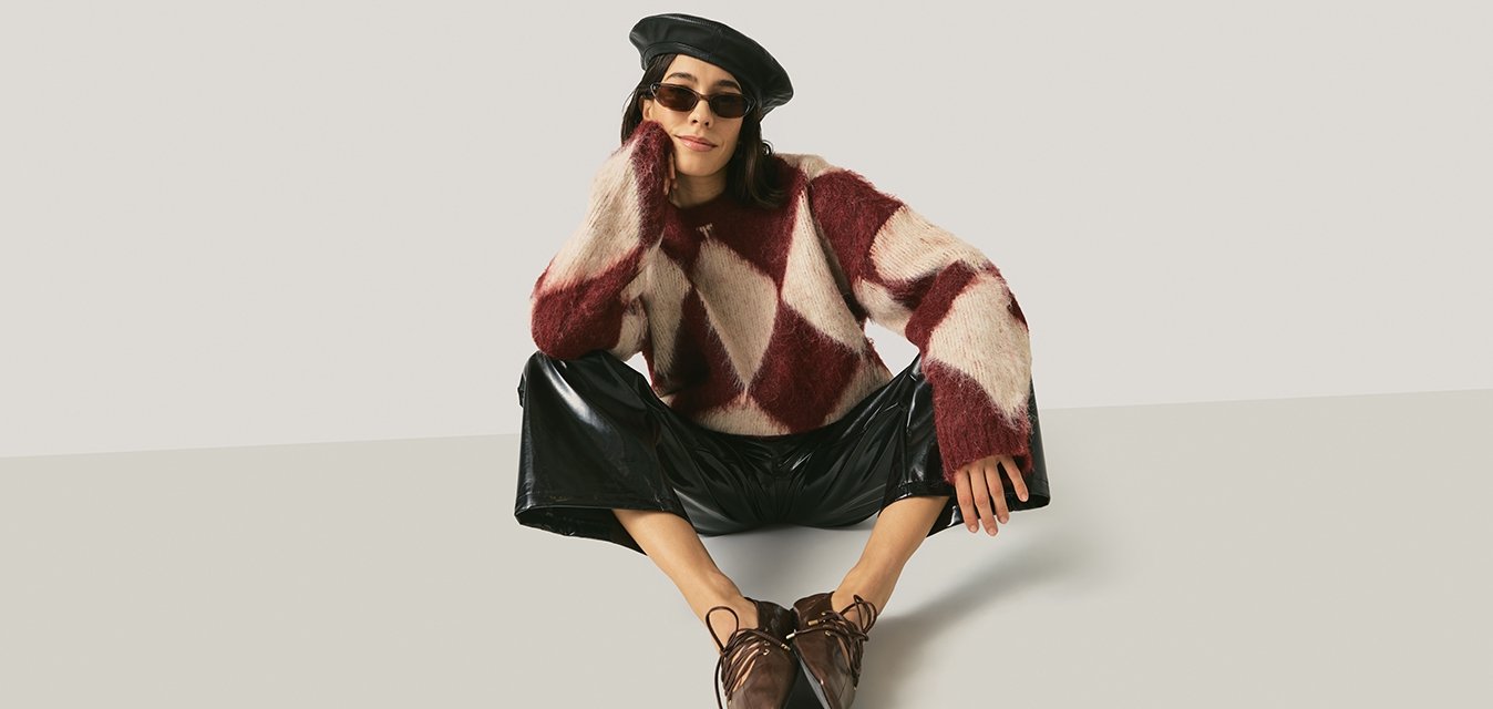 Žena nosící černý baret, sluneční brýle, svetrový vzor v bordó a bílé barvě, černé lesklé kalhoty a hnědé špičaté boty, sedící s roztaženýma nohama.