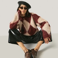 Žena nosící černý baret, sluneční brýle, svetrový vzor v bordó a bílé barvě, černé lesklé kalhoty a hnědé špičaté boty, sedící s roztaženýma nohama.