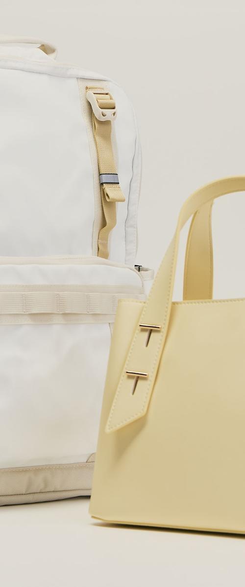 Weißer Rucksack mit beigen Trägern und eine hellgelbe Handtasche mit doppeltem Schnallendetail nebeneinander auf hellem Hintergrund gezeigt.