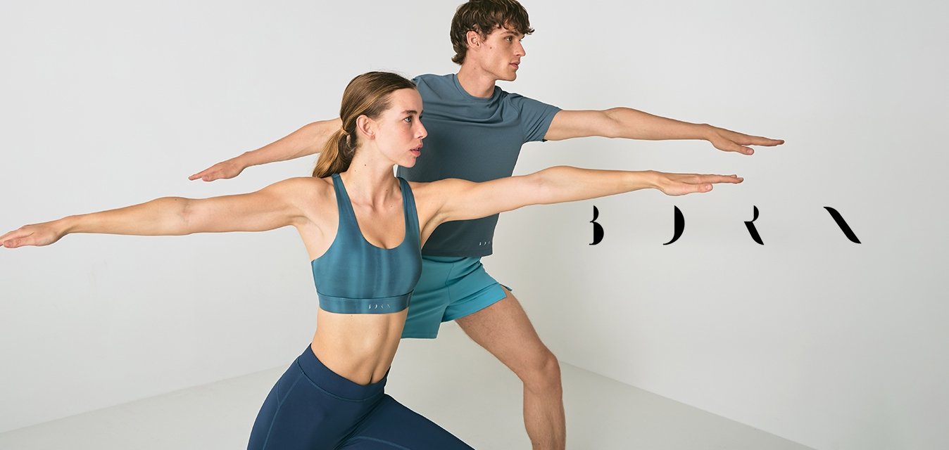 Muž a žena v atletickém oblečení provádějí jogínskou pozici bojovníka, s nataženýma rukama, v minimalistické bílé místnosti.