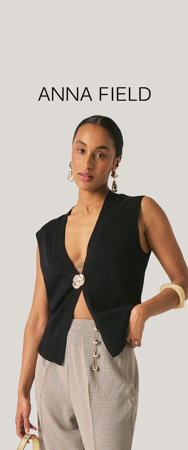 Mujer con chaleco negro sin mangas con un solo botón grande, pantalones de cintura alta con estampado y botones de perla, sosteniendo un bolso pequeño.