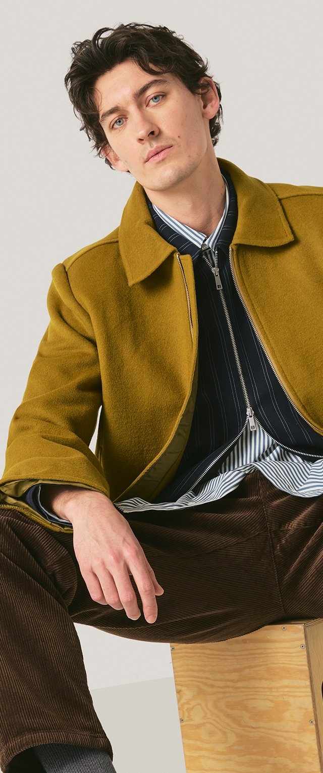 Hombre joven con cabello rizado que lleva un abrigo mostaza, camisa a rayas y pantalones de pana marrón, sentado sobre una caja de madera contra un fondo liso.