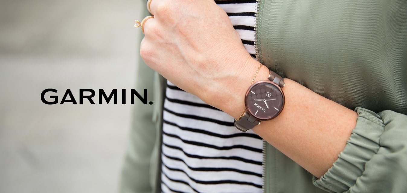 Hnědý kulatý hodinky s tmavou ciferníkem a geometrickými detaily, připevněné na šedém pásku, nošené na zápěstí, vedle pruhované košile a zelené bundy.