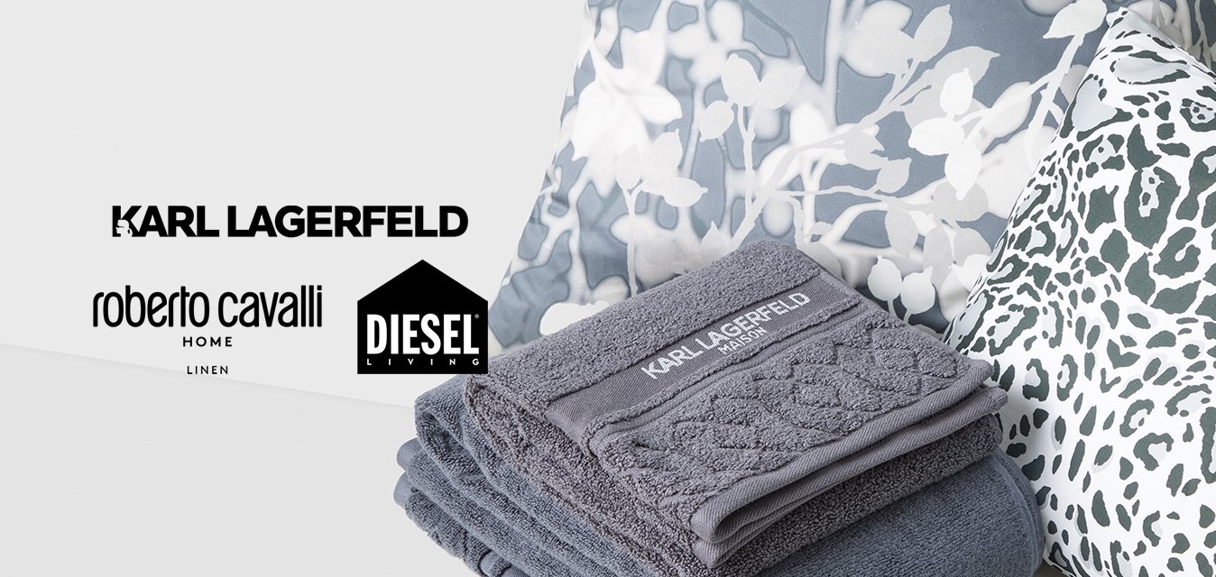 Hromada šedých ručníků Karl Lagerfeld na bílé ploše s polštáři s květinovým a leopardím vzorem v pozadí.