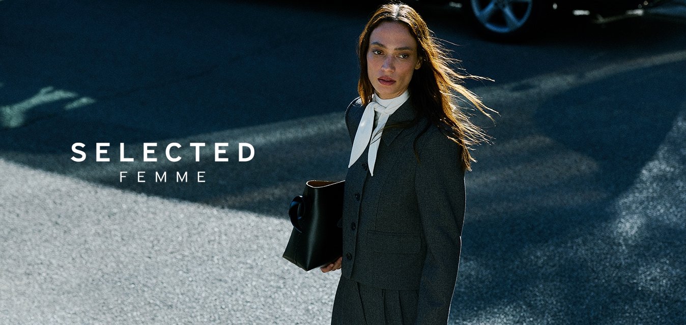 Žena v tmavém obleku a bílém šátku, která nese černou tašku, jde po sluncem osvětlené ulici poblíž zeleno-bílé značky a zaparkovaného auta.