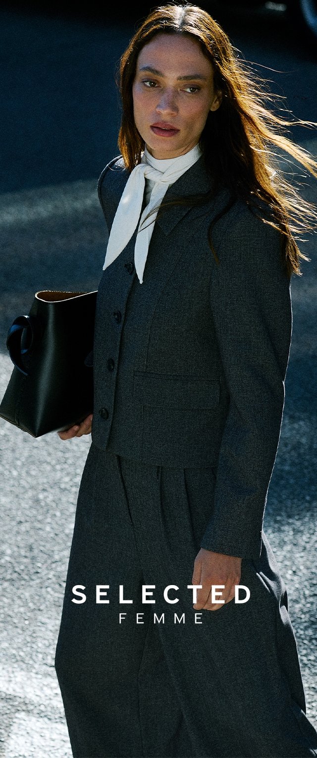 Vrouw met lang bruin haar, gekleed in een donkere maatpak, met een witte sjaal om de nek en een zwarte handtas, die in de zon buiten wandelt.