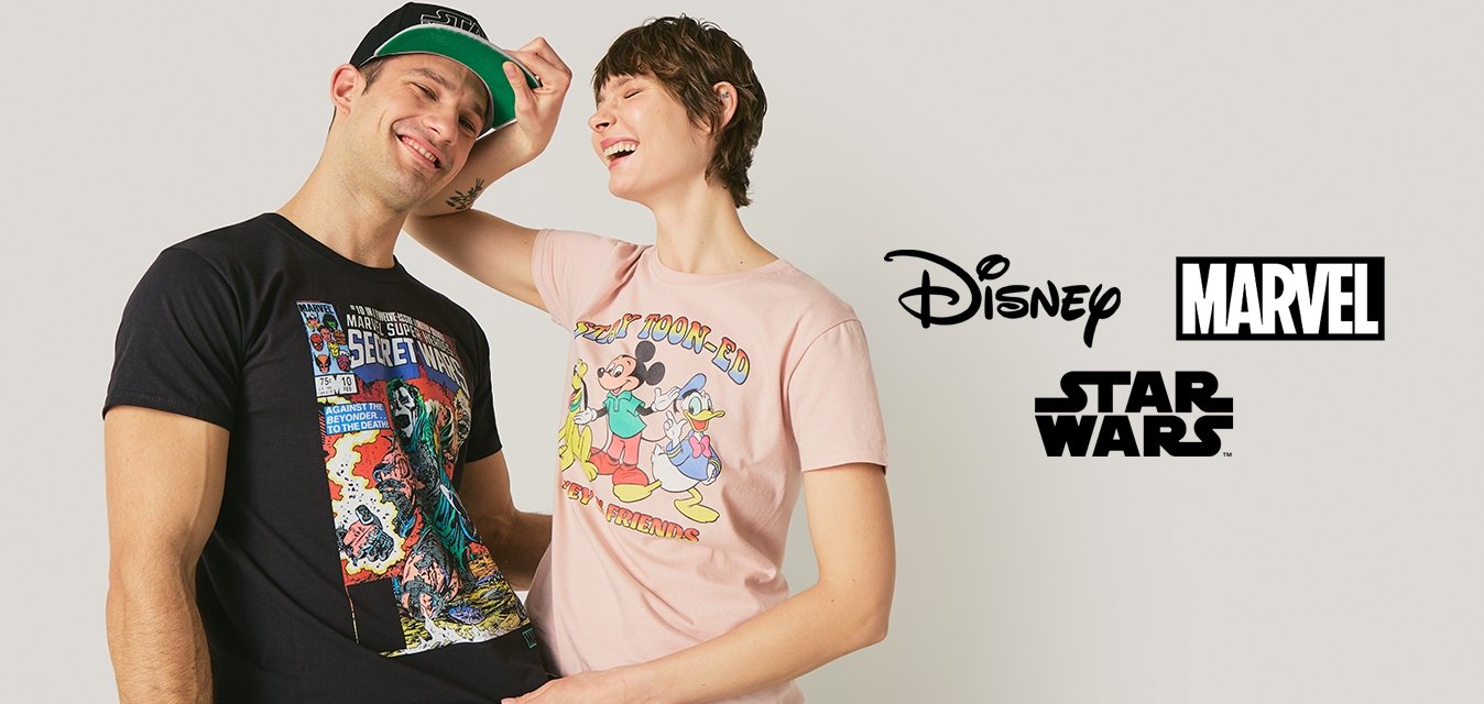 Dva mladí dospělí se usmívají, jeden má na sobě černé tričko a čepici s komiksovým motivem, druhý si upravuje čepici a nosí růžové tričko s kreslenou postavičkou.