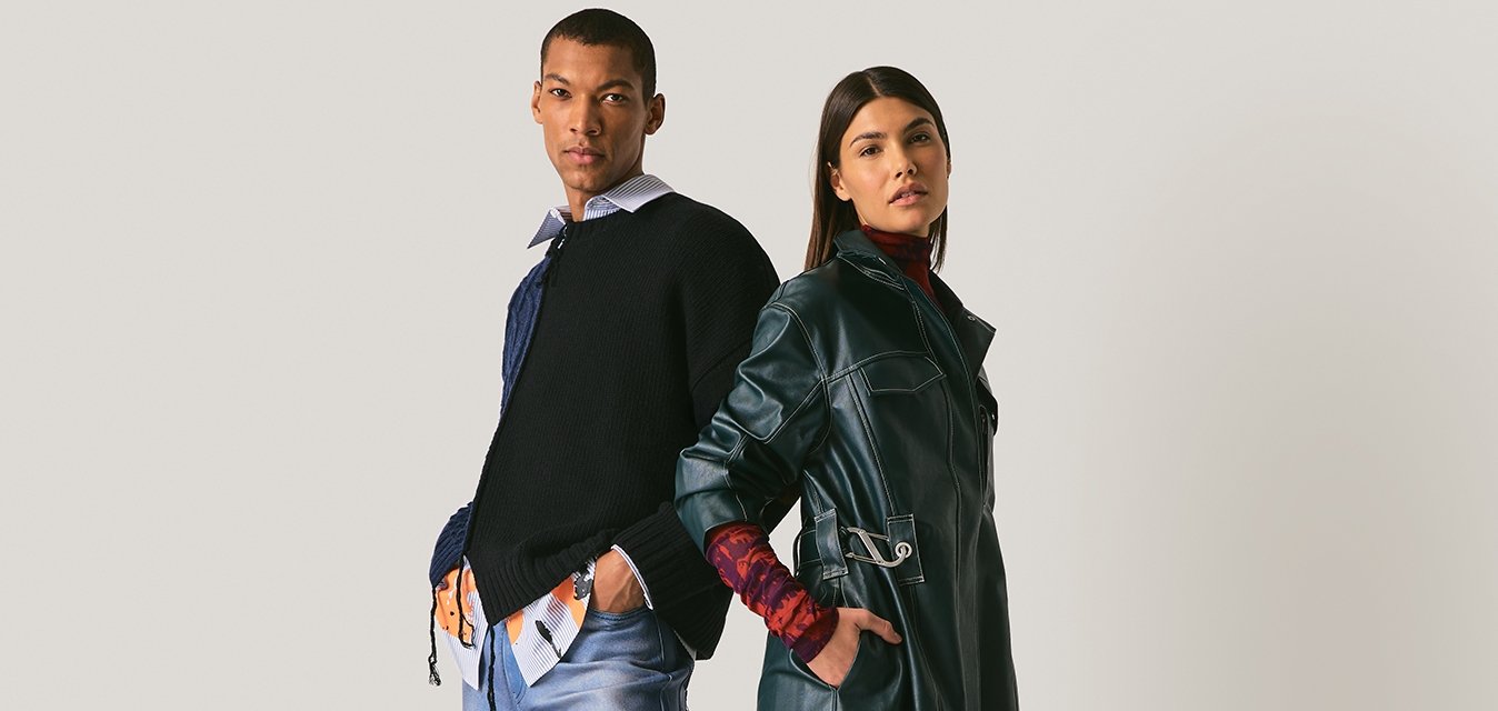 Muž a žena stojící zády k sobě, oblečeni v moderních vícevrstvých outfitech s denimu, koženým trenčkotem a pohodlnou obuví na neutrálním pozadí.