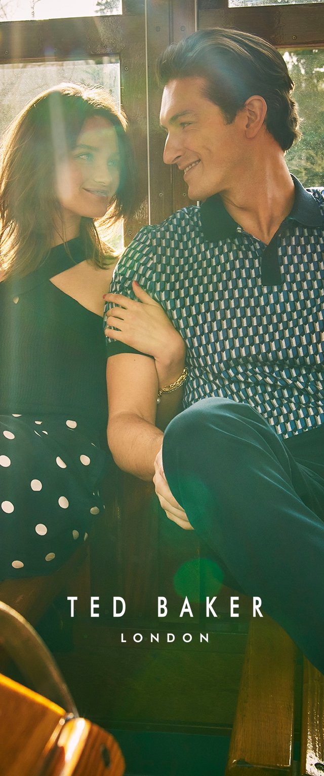 Modelo con un vestido negro con cortes y lunares blancos, junto a un hombre con una camiseta de polo de estampado geométrico azul, sentados en un entorno de madera.