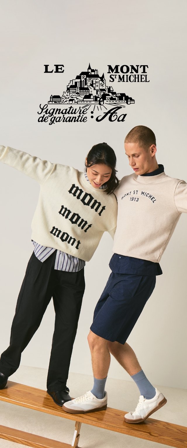Twee mensen balanceren op een houten bankje, gekleed in truien met de tekst "Mont St Michel", voor een muur met een afbeelding en opschrift van Mont St Michel.