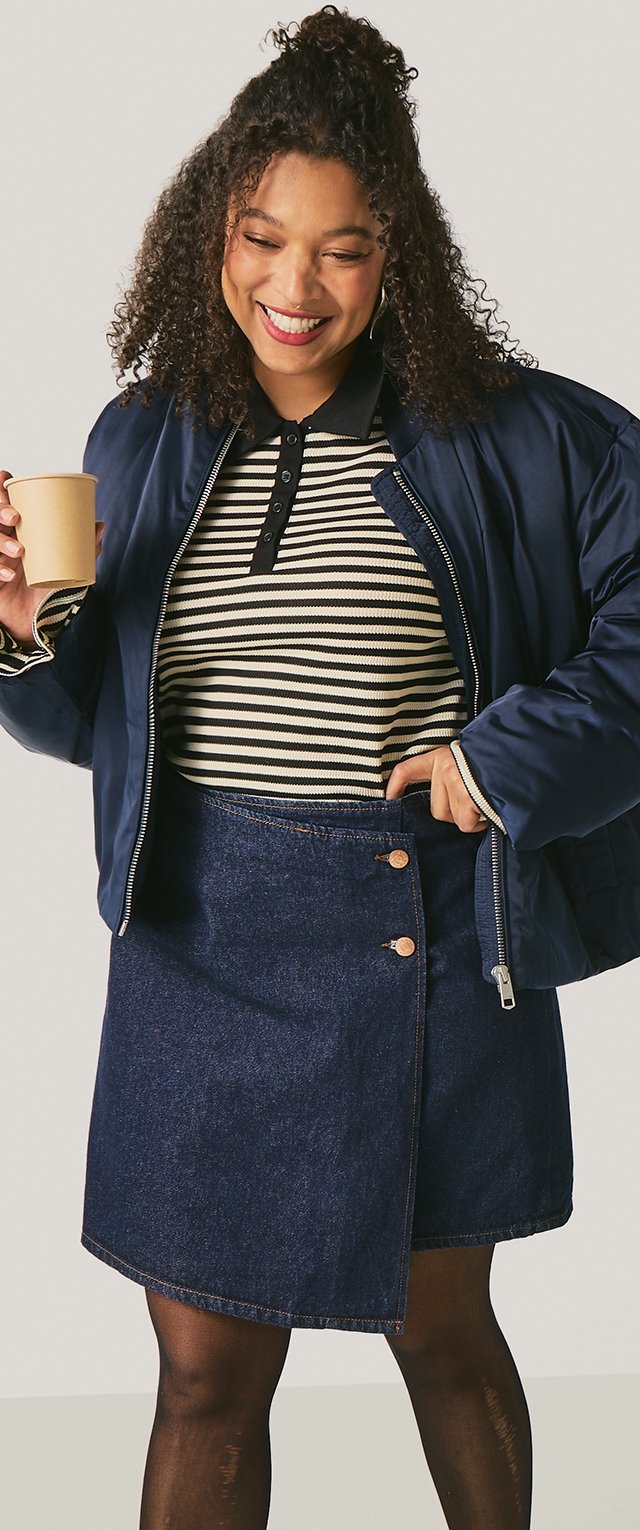 Mujer sonriente con cabello rizado que lleva una chaqueta bomber azul marino, camiseta tipo polo a rayas en blanco y negro, falda de mezclilla y sostiene un vaso de papel en su mano izquierda.