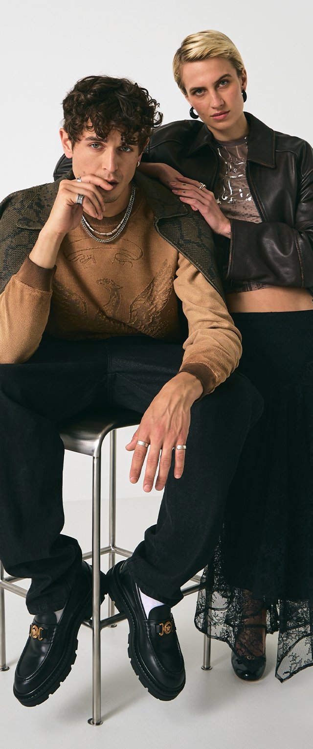 Zwarte loafers met gouden hardware, een tan kleurige trui met reliëfdetails, en een bruin leren jack over een doorzichtig topje en zwarte kanten rok.