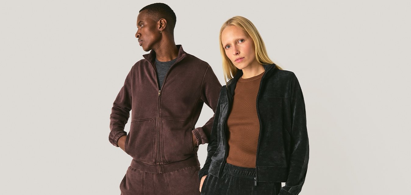 Dva modelové stojící vedle sebe, oblečeni v neformálních sametových teplákových soupravách, jeden v tmavě hnědé a druhý v černé, na jednobarevném pozadí.