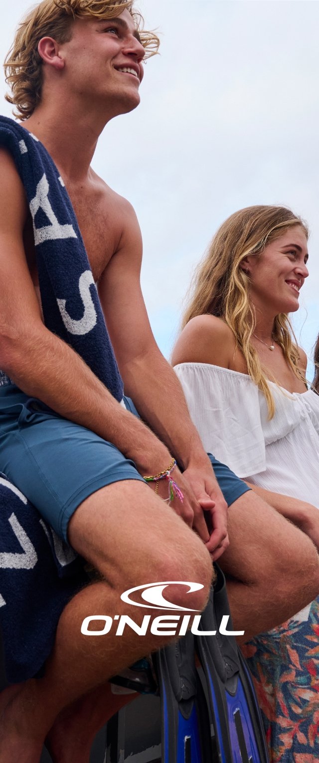 Deux personnes sont assises dans un bateau. L'homme porte un short de bain bleu et tient une serviette. La femme porte un top blanc à épaules dénudées et un short floral.