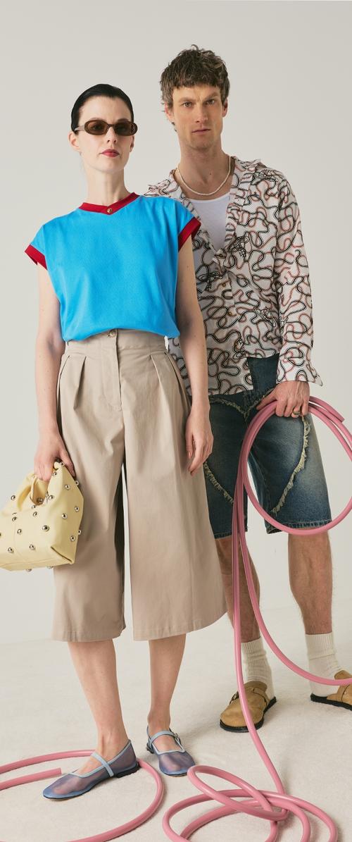 Frau in blauem Top, beigen Culottes, Sonnenbrille, hält eine gelbe Tasche mit Nieten; Mann in gemustertem Hemd, Denim-Shorts, Sandalen hält einen pinken Schlauch.
