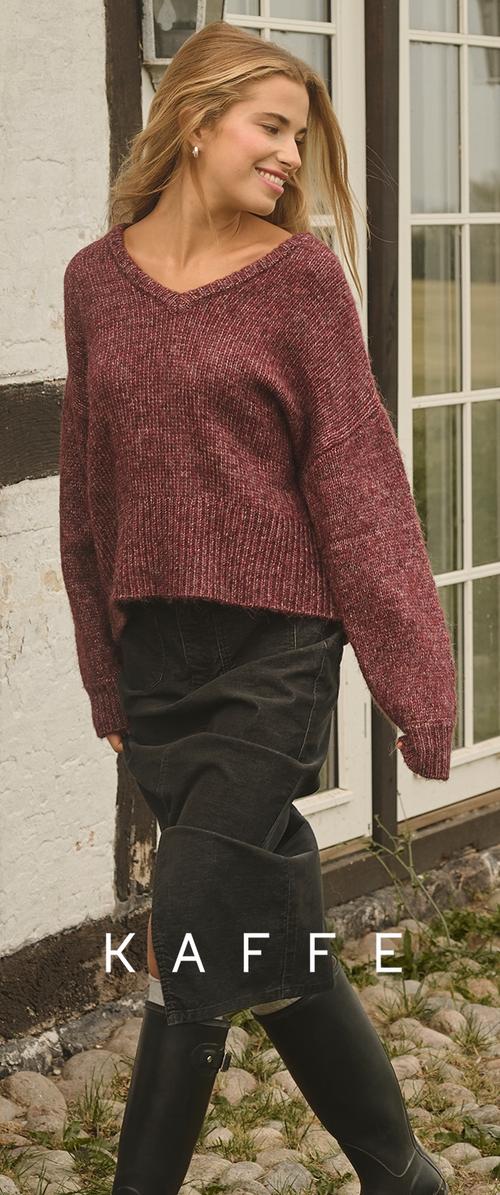 Kvinde iført en bordeaux strikket sweater, sort nederdel og sorte støvler står udendørs nær en rustik hvid- og sortmalet bindingsværksbygning.