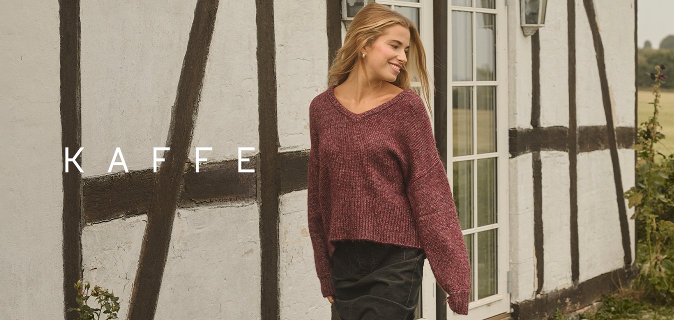 Mladá žena v vínovém svetru a černých botách kráčí po kostkované cestě vedle chalupy se střechou z došek a bílými květinami v blízkosti.